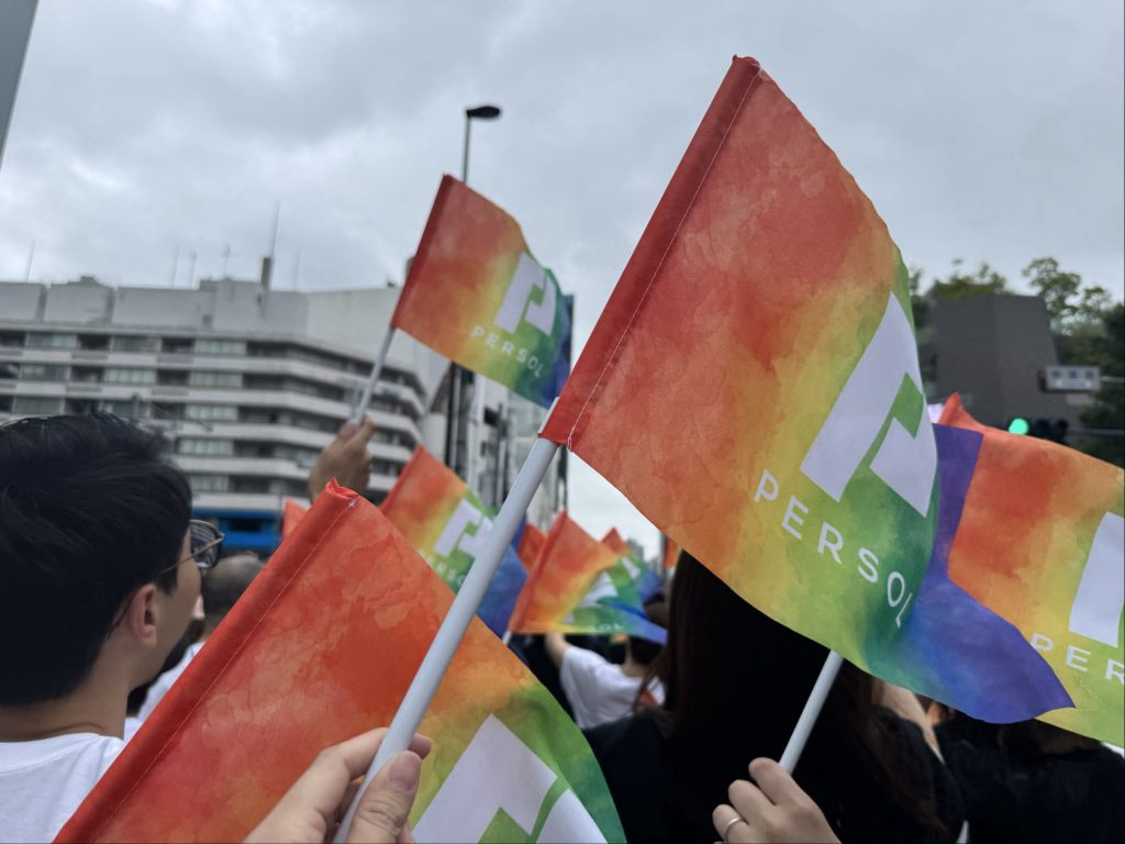 「Tokyo Pride」参加の様子。屋外で人々が虹色の旗を掲げながら行進している様子。周囲にはビルが立ち並び、空が広がっている。